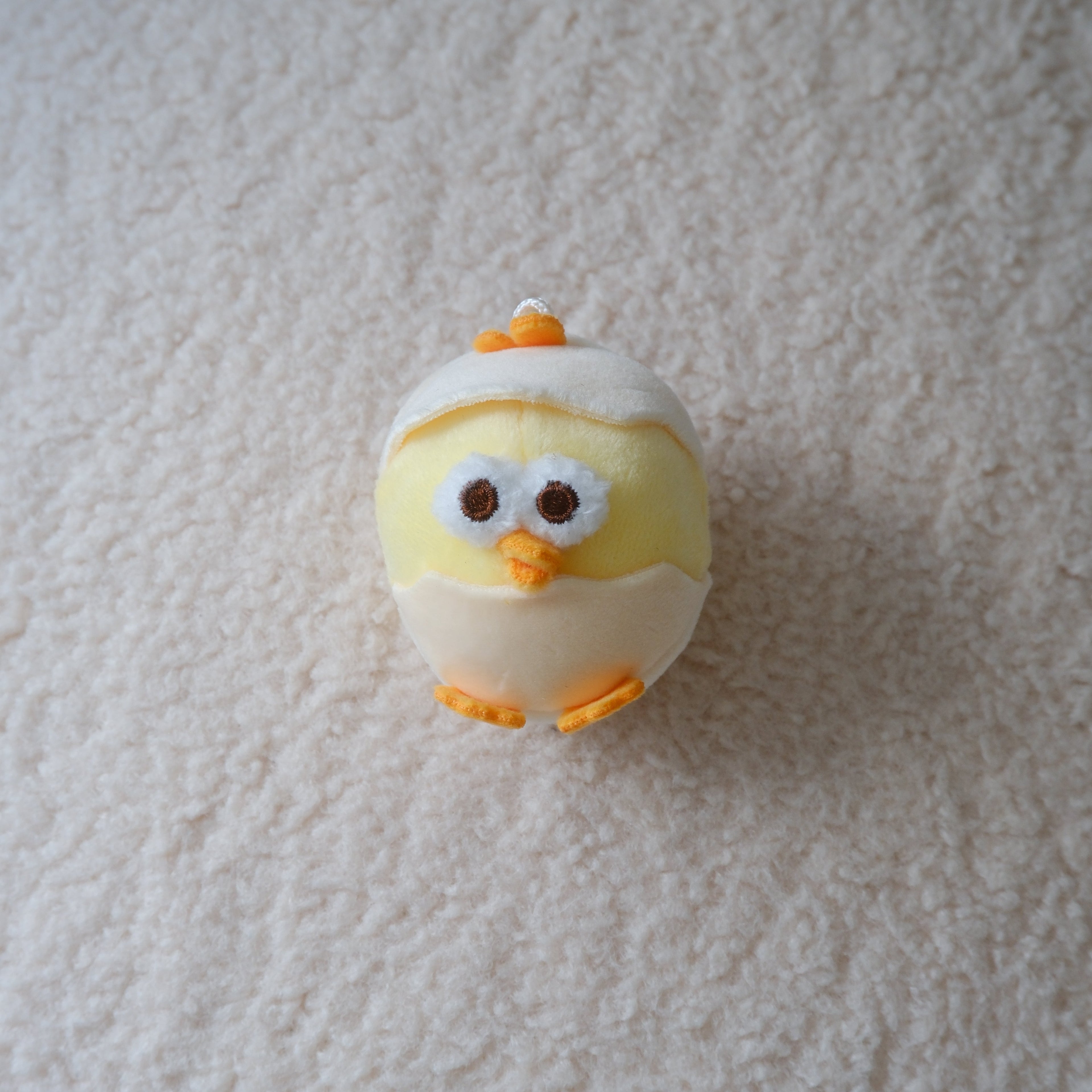 A small plush chick toy wearing a white eggshell, standing on a textured off-white carpet. The chick has yellow fabric body, orange beak and feet, and large embroidered eyes.