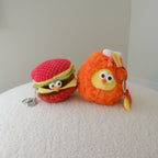 Two small plush keychains resting on a textured cream surface against a plain light gray wall. One keychain is shaped like a red crocheted hamburger with green and yellow details, and the other is an orange fuzzy drumstick with a yellow face and small french fry keychain.