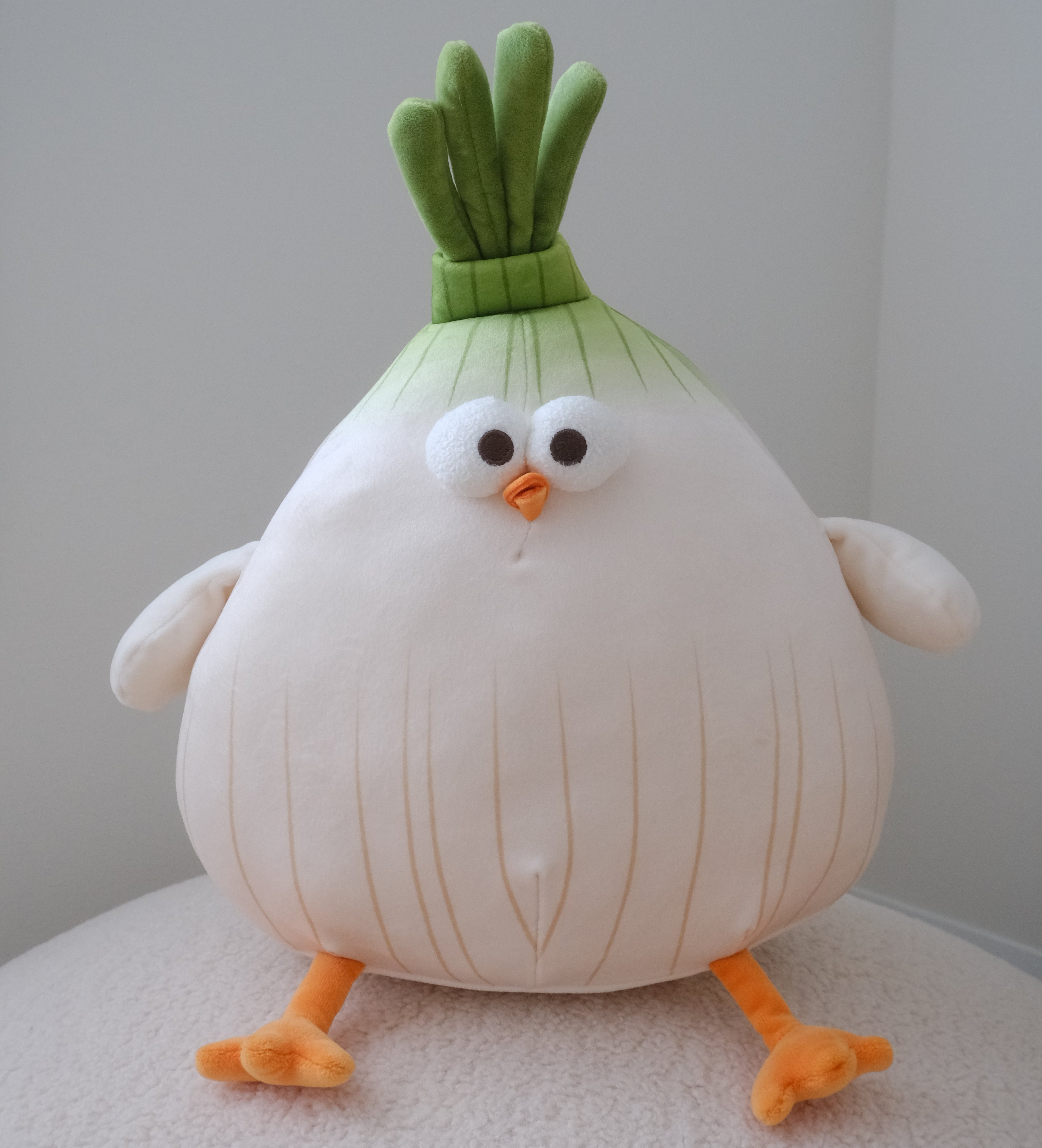A cute, round plush toy designed like a cartoon onion character, featuring a large white body with vertical yellowish lines, green leafy top, orange beak and feet, and small white wings, sitting on a textured white surface against a neutral gray background.