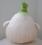 A plush onion-shaped pillow with a cream-colored body, light green stem, and small side ears, sitting on a textured cream surface against a neutral gray wall.