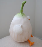 A side profile of a plush toy shaped like a stylized onion with a green stem, white bulbous body, small white eye, and orange feet, photographed indoors on a light surface against a plain wall.