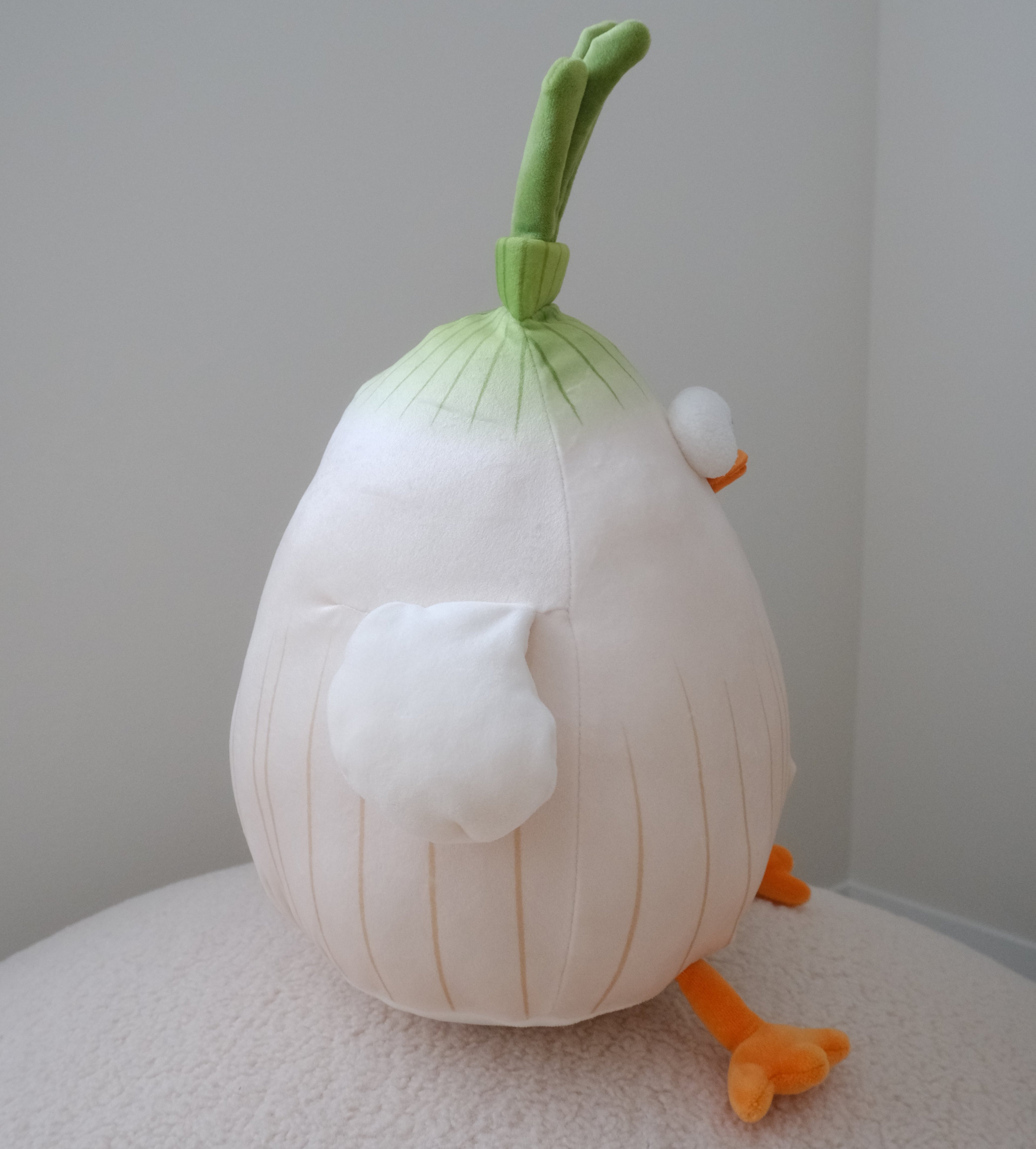 A side profile of a plush toy shaped like a stylized onion with a green stem, white bulbous body, small white eye, and orange feet, photographed indoors on a light surface against a plain wall.