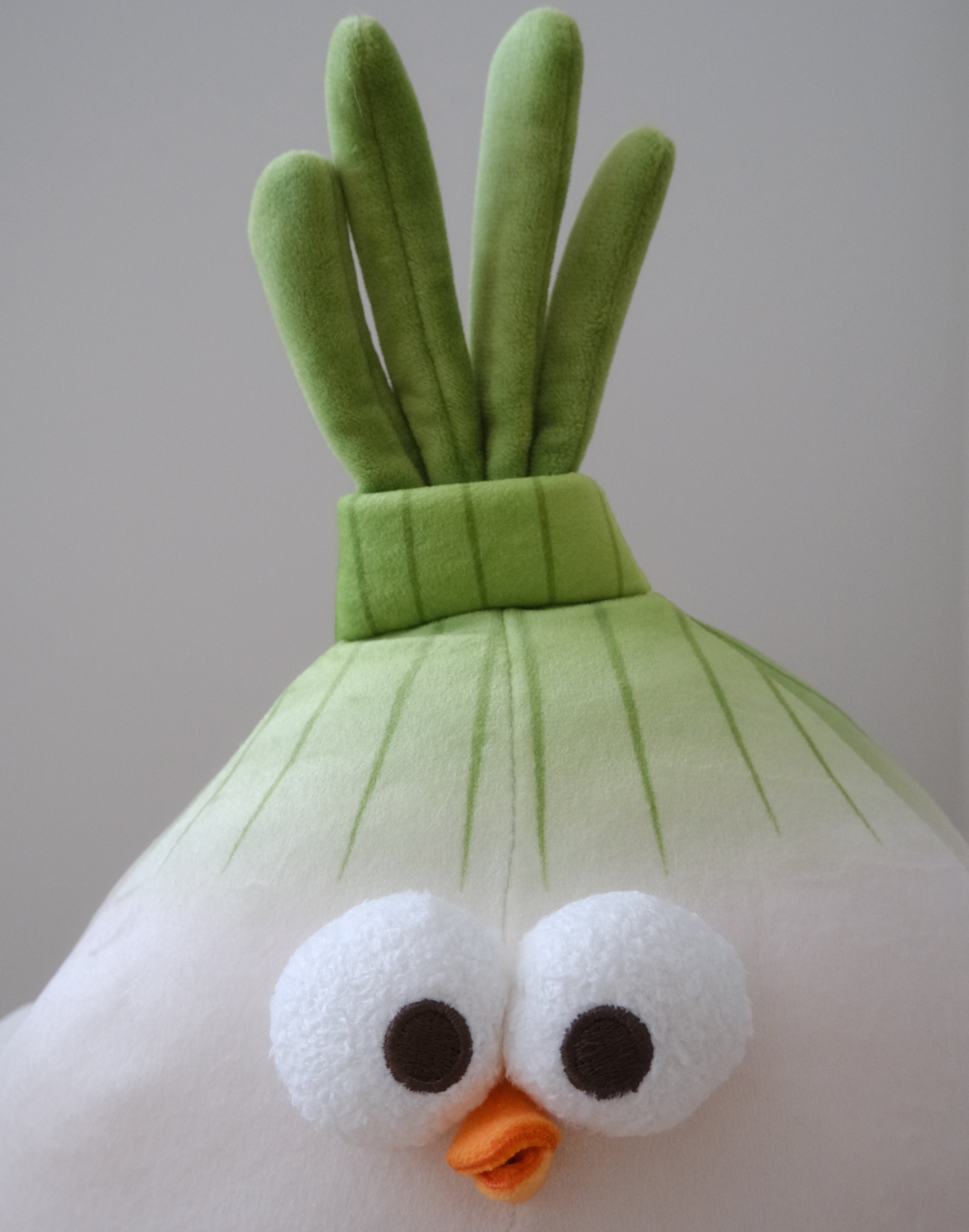 A plush toy shaped like a stylized onion character, with a white body, green leafy top, large white eyes, and a small orange beak, photographed against a plain light gray background.