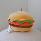 A plush round toy shaped like a hamburger, with a tan bun, green lettuce, red patty, and brown cheese layers, sitting on a textured cream surface against a plain light gray wall.