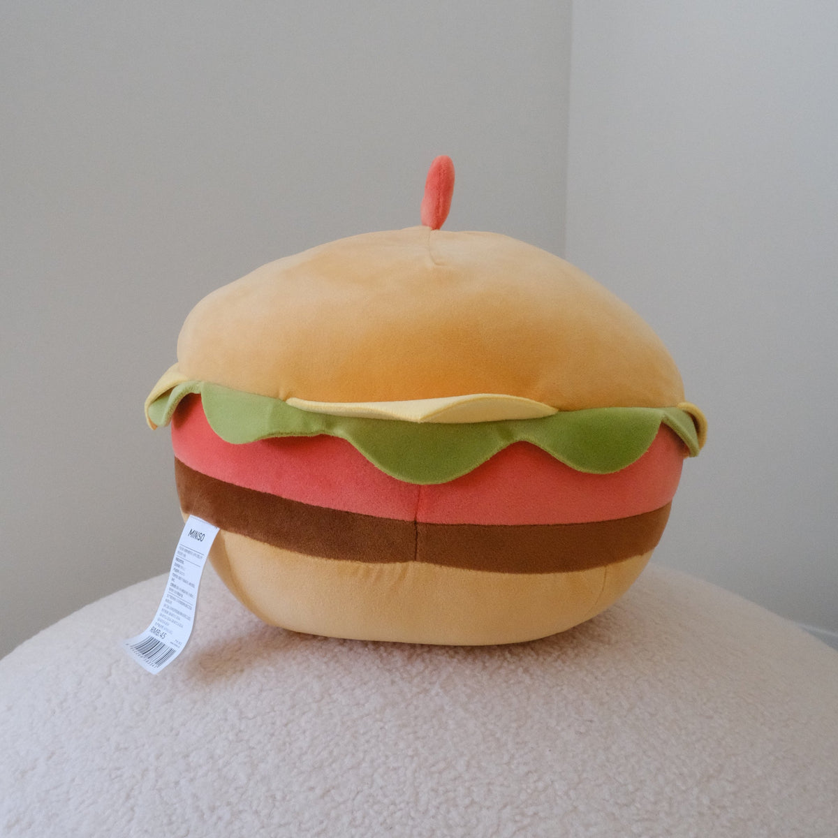 A plush round toy shaped like a hamburger, with a tan bun, green lettuce, red patty, and brown cheese layers, sitting on a textured cream surface against a plain light gray wall.
