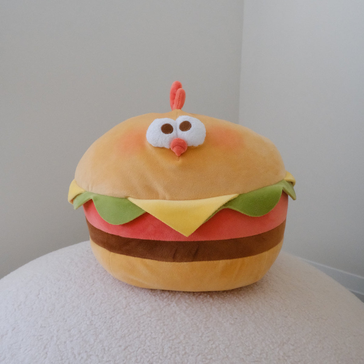 A plush toy shaped like a chicken burger with a cute face, sitting on a textured white surface against a plain light gray background.