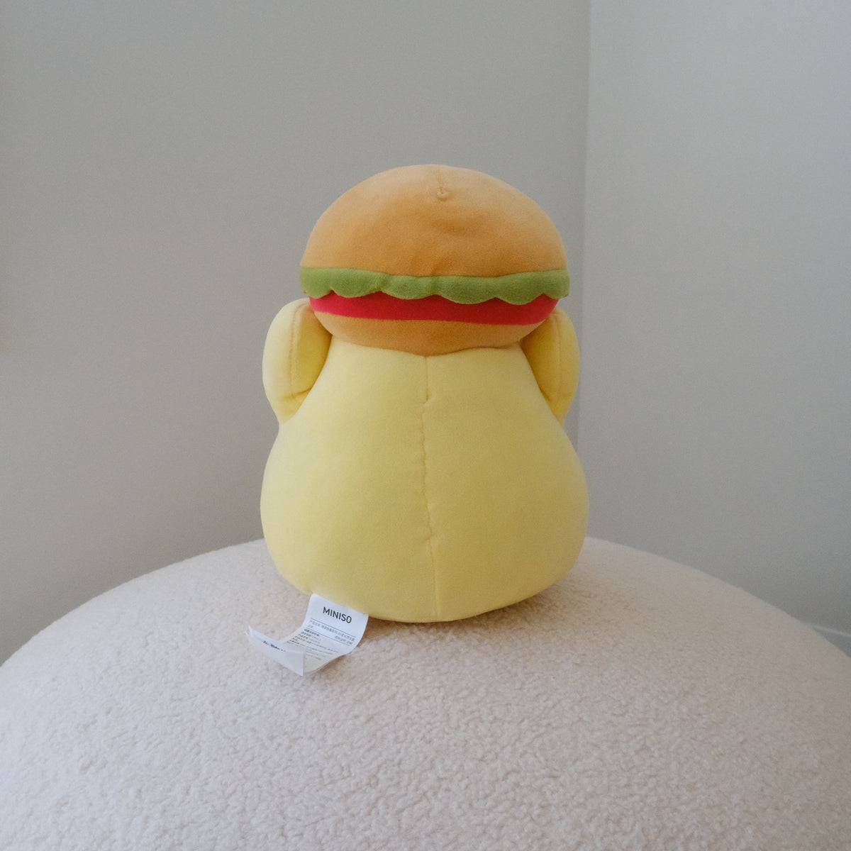 A yellow plush chicken toy shaped wearing a hamburger hat, sitting on a textured cream-colored surface against a plain light gray wall.