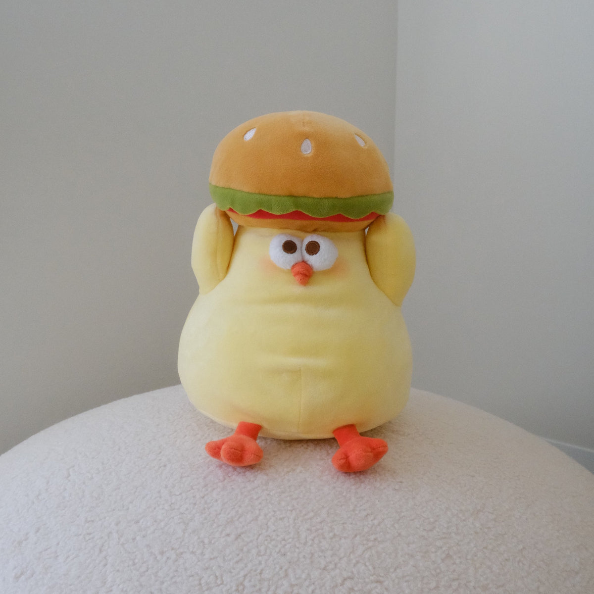 A yellow plush toy shaped like a cute chicken wearing a hamburger hat, sitting on a textured white surface against a plain light gray wall.