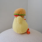 A yellow plush toy chicken wearing a hamburger hat, sitting on a textured white surface against a plain light gray wall.