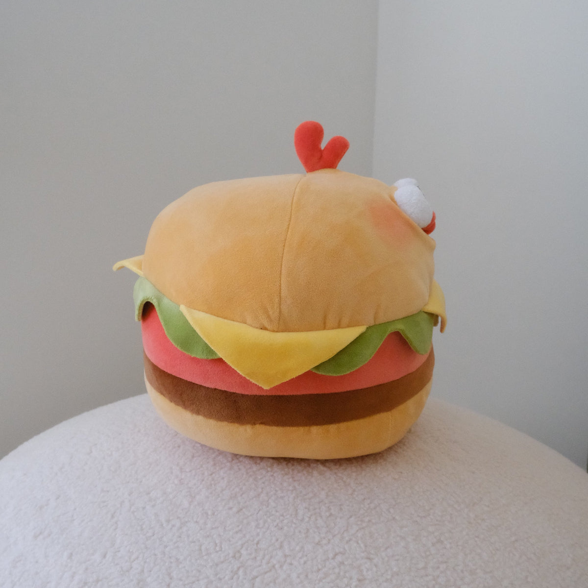 A plush toy shaped like a hamburger with a small red antler on top and a white eye detail on the side, sitting on a textured white surface against a plain light gray wall.