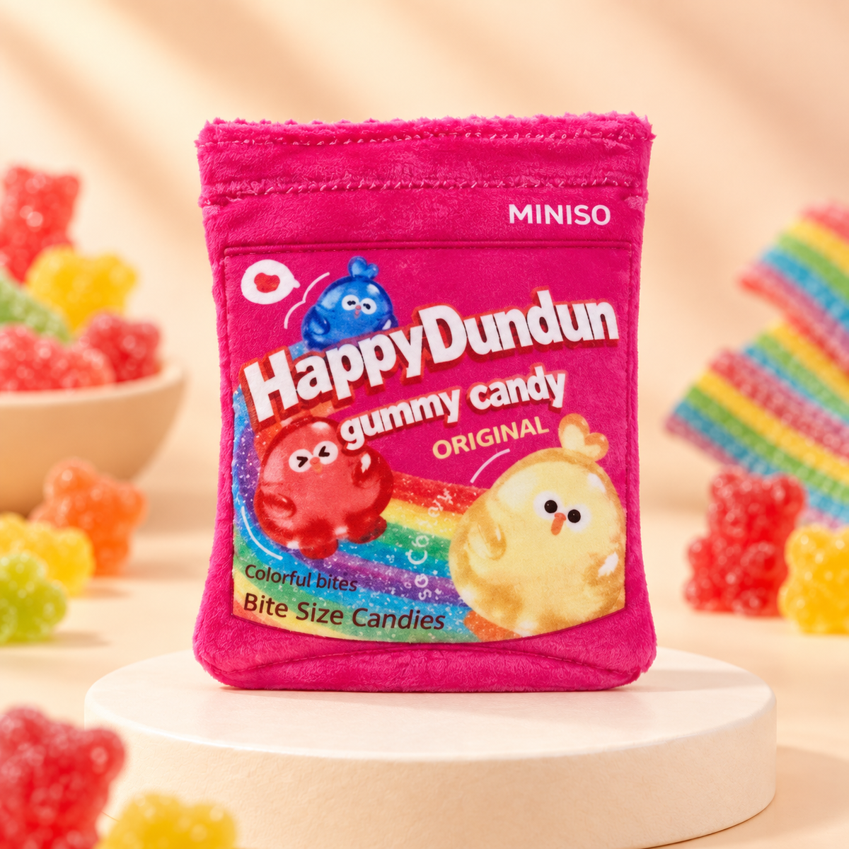 A novelty felt packaging wrapper for gummy candy stands upright on a white round pedestal. The wrapper is magenta pink with rainbow graphics, and the text 'HappyDundun gummy candy original" and "Colorful bites Bite Size Candies"