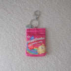A small pink keychain pouch designed to look like a candy package, featuring a cartoon gummy chicken and the text 'Happy Dundun Candy', with silver metal key rings attached, placed on a textured off-white surface.