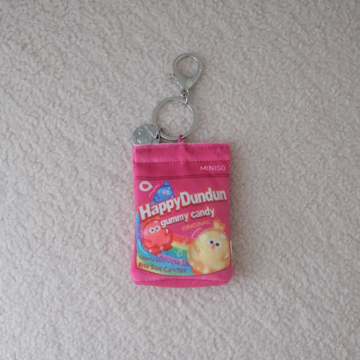 A small pink keychain pouch designed to look like a candy package, featuring a cartoon gummy chicken and the text 'Happy Dundun Candy', with silver metal key rings attached, placed on a textured off-white surface.
