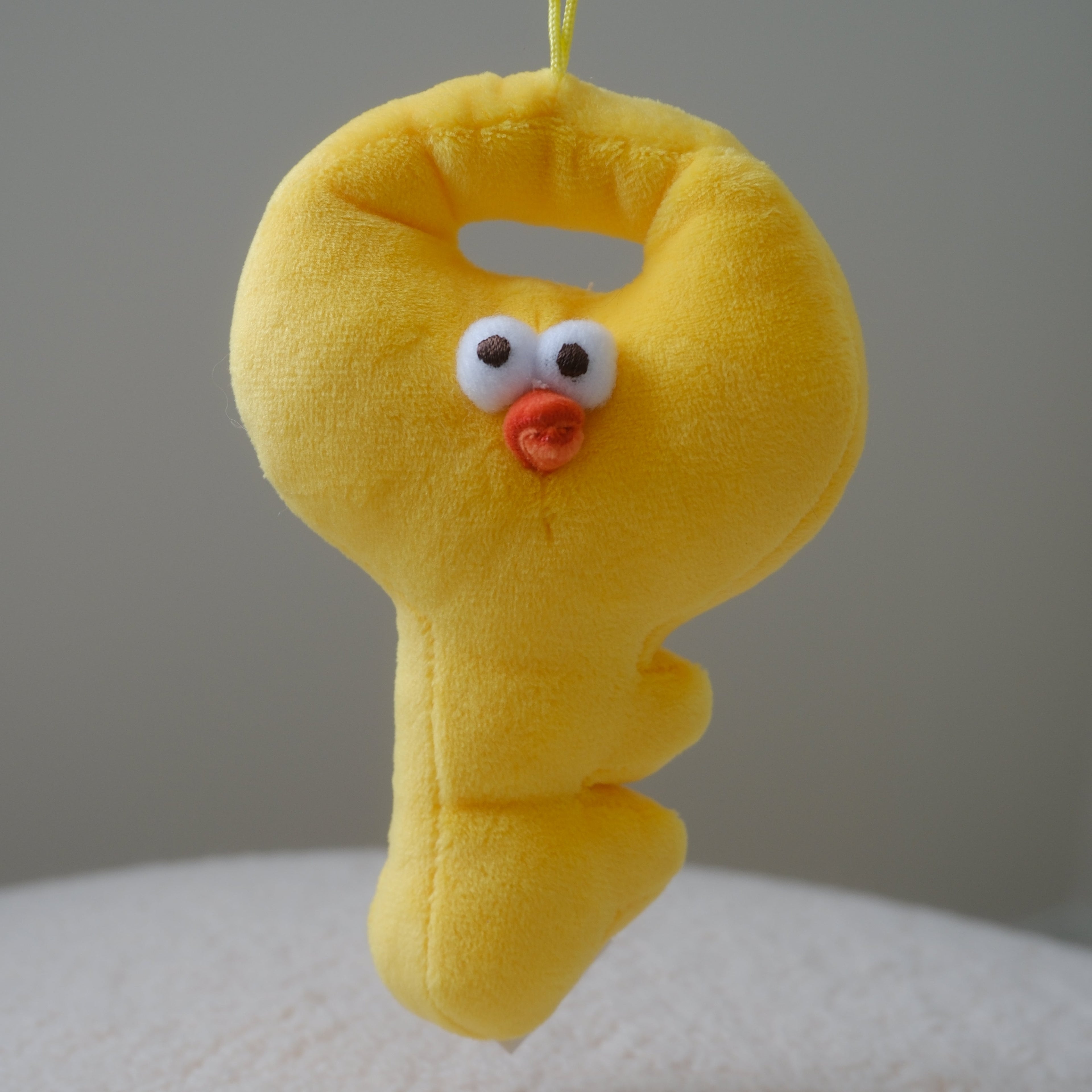 A yellow plush key-shaped chicken with a cute face, hanging from a yellow plastic clip against a neutral gray background.