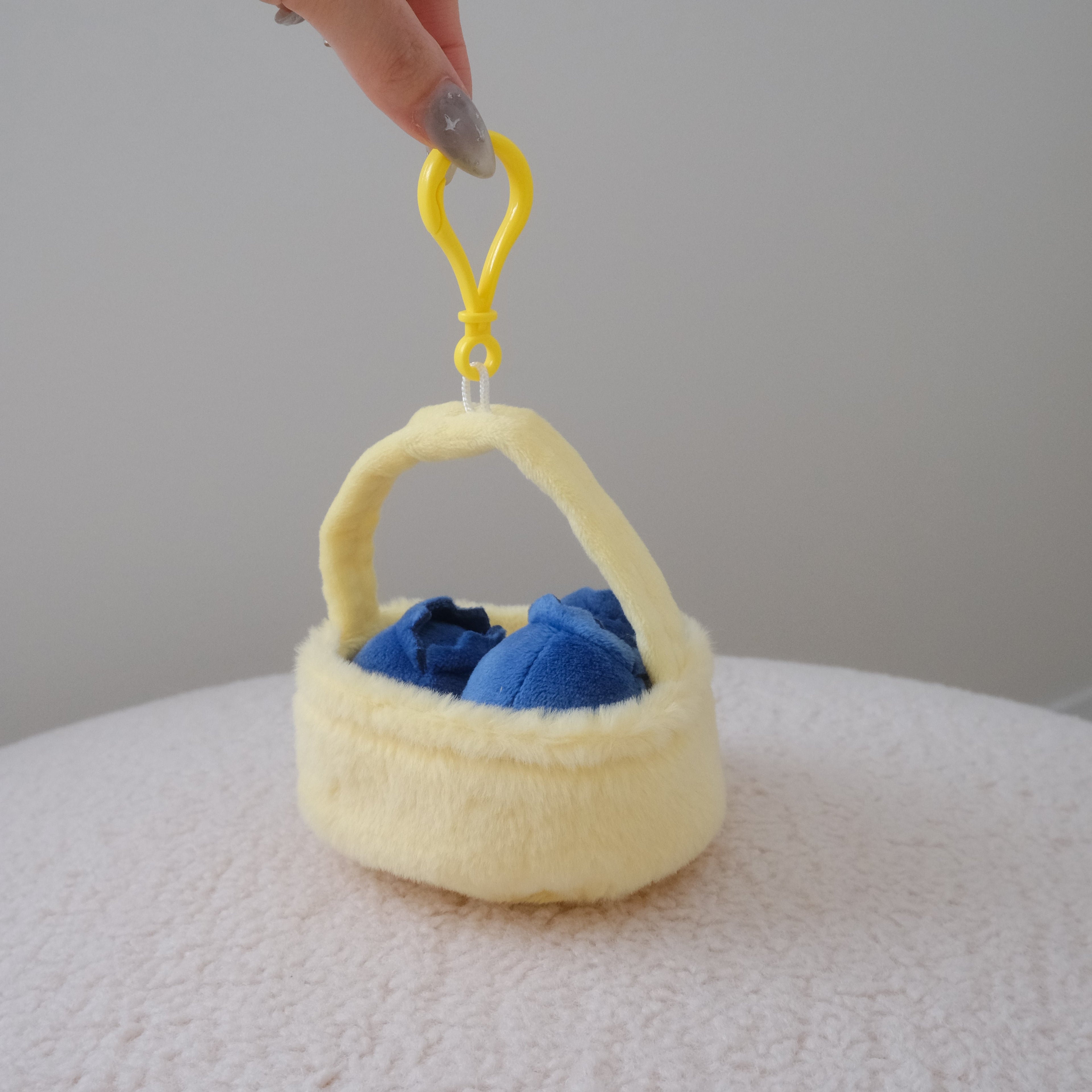 A hand holding a small plush basket bag in pale yellow with a bright yellow clip, containing three plush blueberries, set against a neutral light gray background.