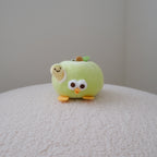 A small plush toy shaped like a light green apple with cartoon eyes, a smiling face, and orange feet, sitting on a textured cream surface against a plain light gray wall.