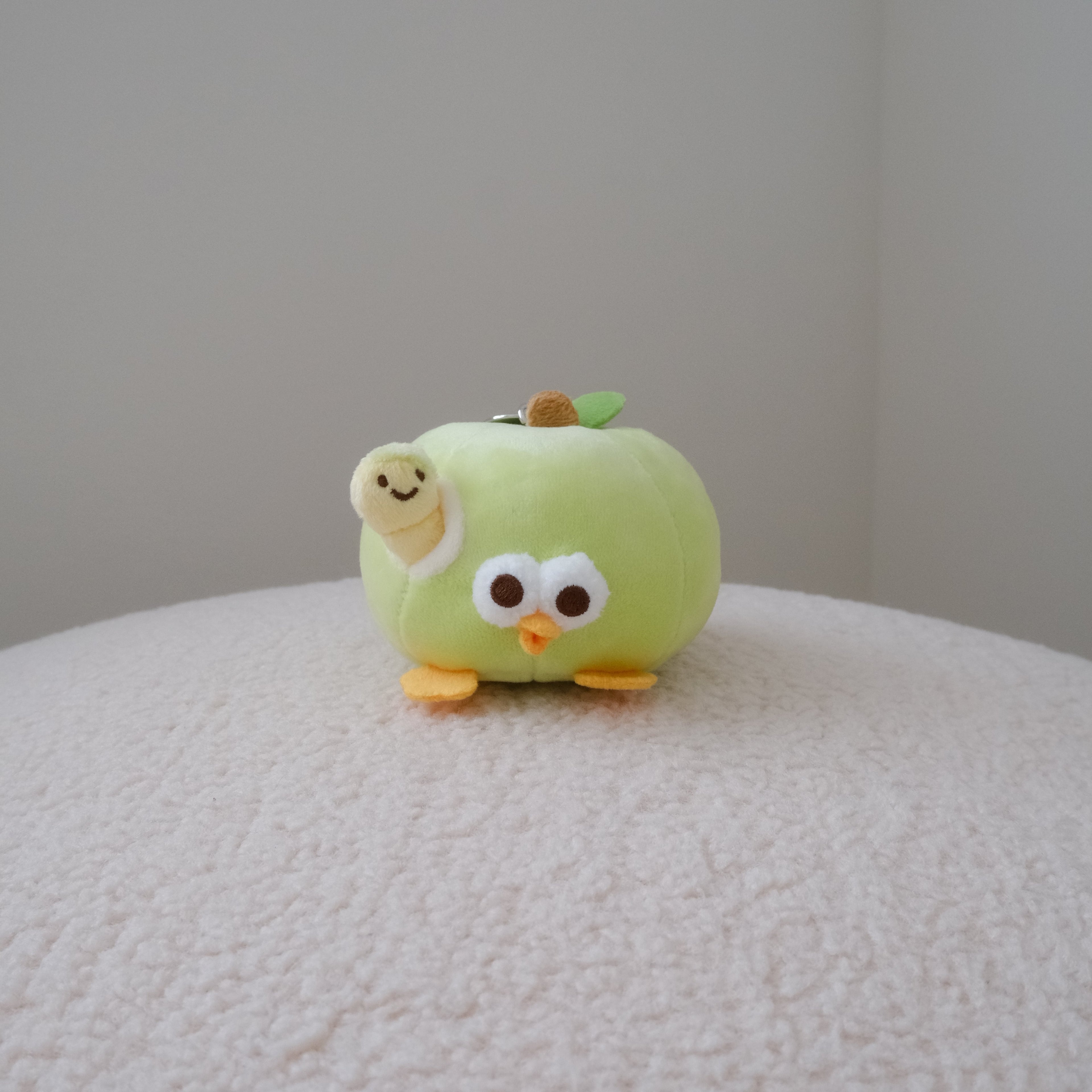 A small plush toy shaped like a light green apple with cartoon eyes, a smiling face, and orange feet, sitting on a textured cream surface against a plain light gray wall.