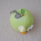 A small plush keychain shaped like a light green apple with a yellow stem, white and yellow face details, and a silver metal key ring, photographed on a soft beige textured fabric background.