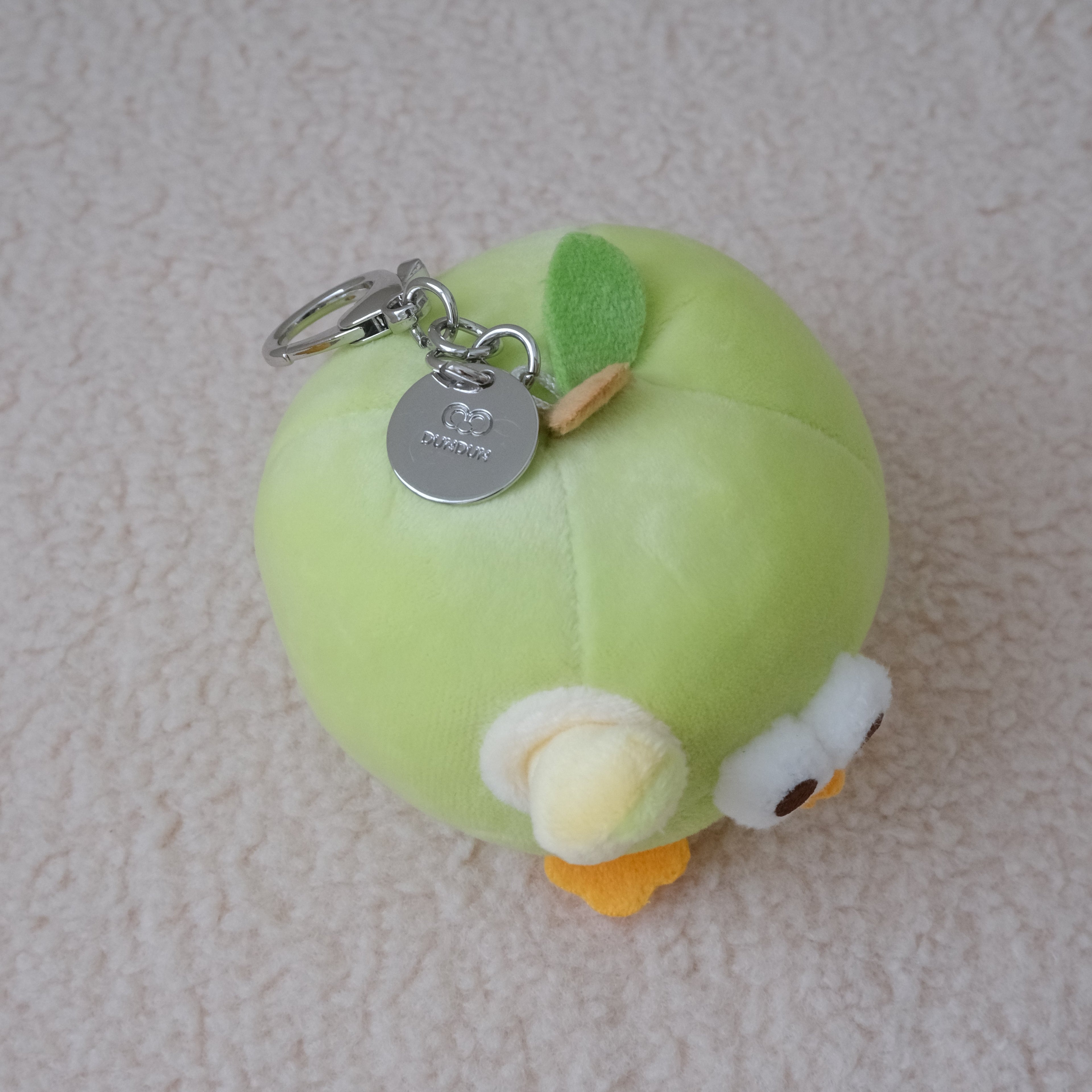 A small plush keychain shaped like a light green apple with a yellow stem, white and yellow face details, and a silver metal key ring, photographed on a soft beige textured fabric background.