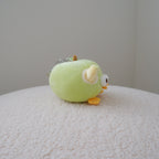 A small plush toy shaped like a round green apple with yellow-orange feet and a white eye detail, sitting on a textured cream surface against a plain light gray wall.