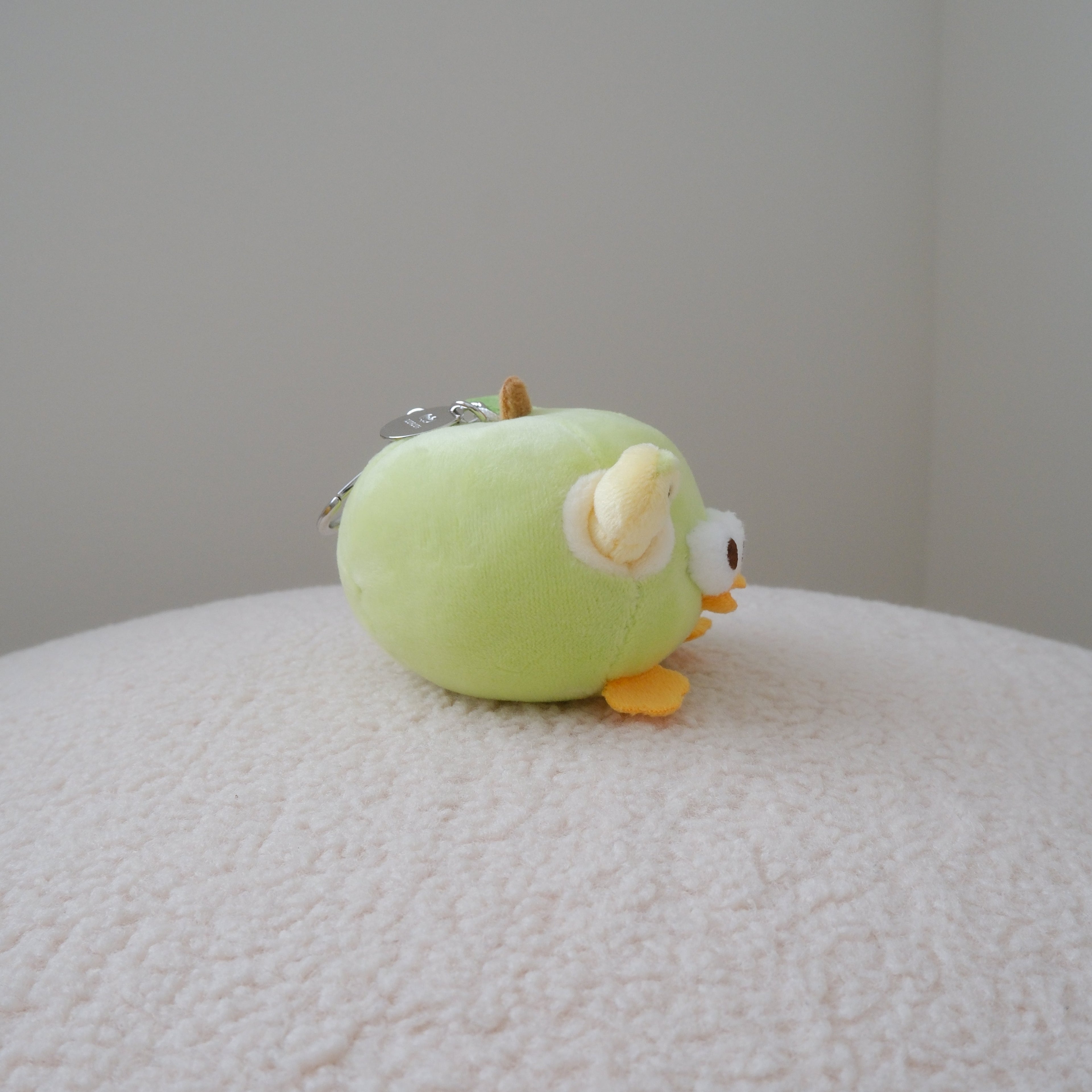A small plush toy shaped like a round green apple with yellow-orange feet and a white eye detail, sitting on a textured cream surface against a plain light gray wall.