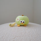 A small green apple plush character toy with large white eyes, orange feet, and a yellow worm attached to its side, sitting on a textured white surface against a plain light gray wall.