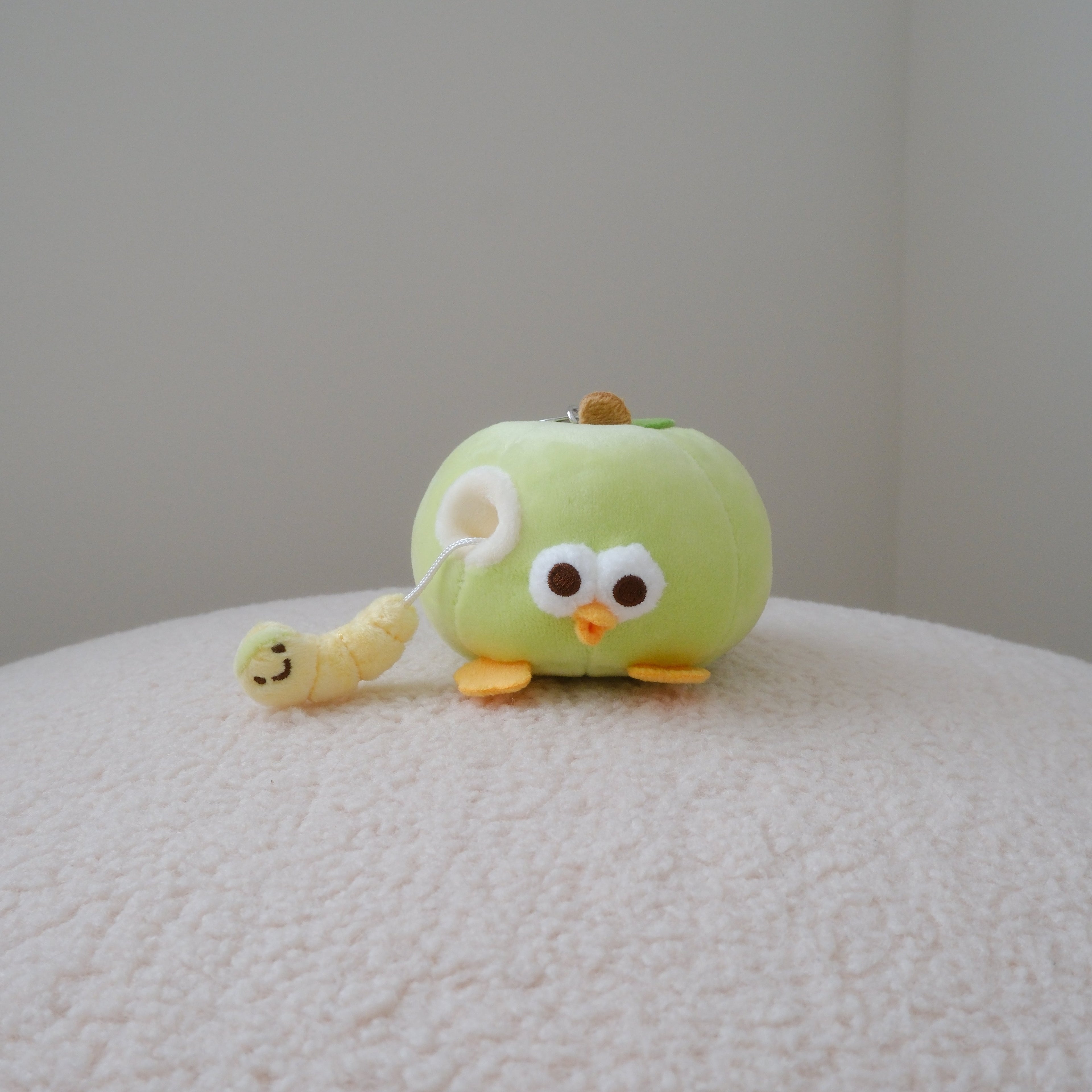 A small green apple plush character toy with large white eyes, orange feet, and a yellow worm attached to its side, sitting on a textured white surface against a plain light gray wall.
