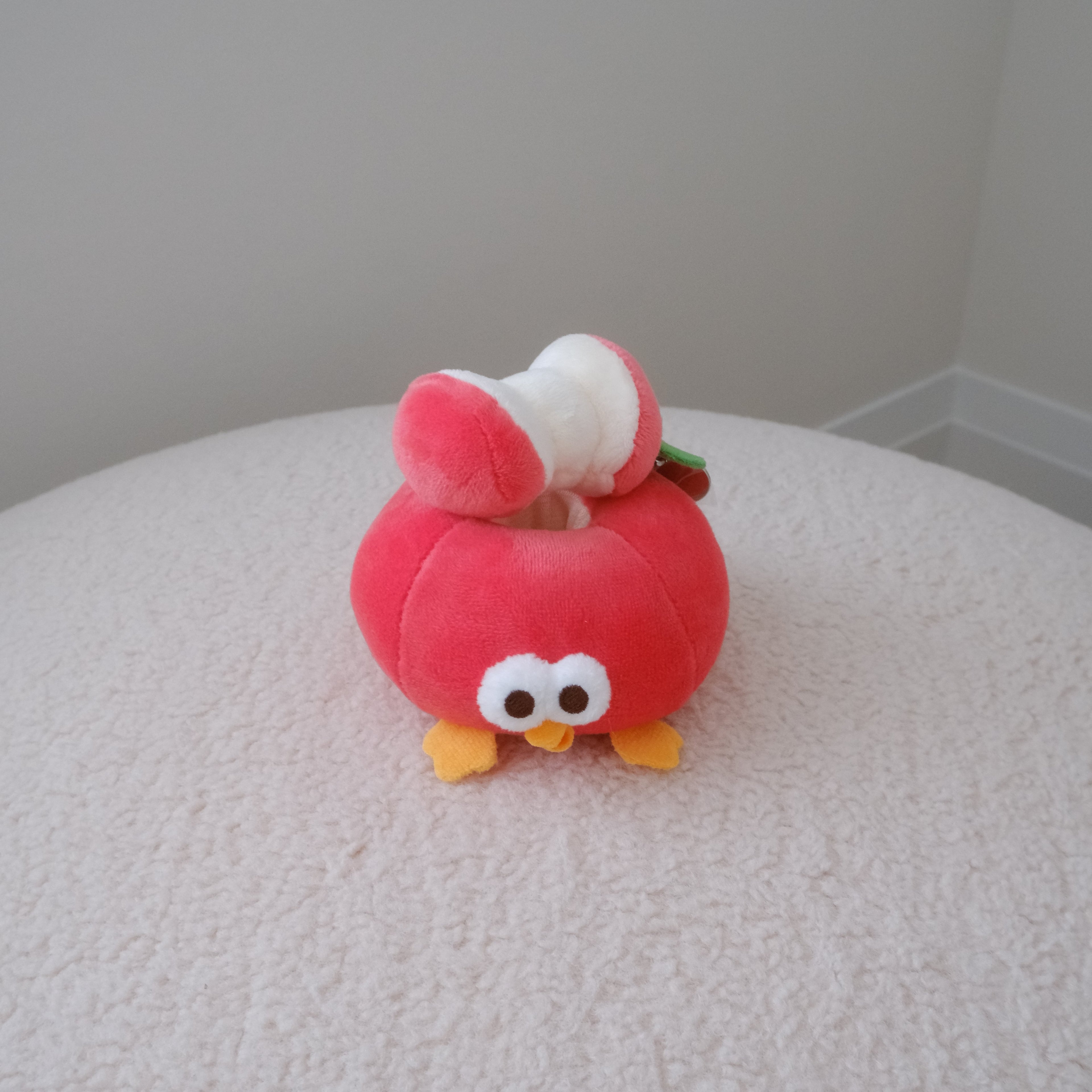 A small red plush toy shaped like an apple with large white eyes, yellow feet, and an apple core on its head, sitting on a textured white surface against a plain light gray wall.