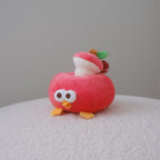 A small plush toy shaped like a red apple with a cute bird face, yellow feet, and a mushroom on top, sitting on a textured white surface against a plain gray background.