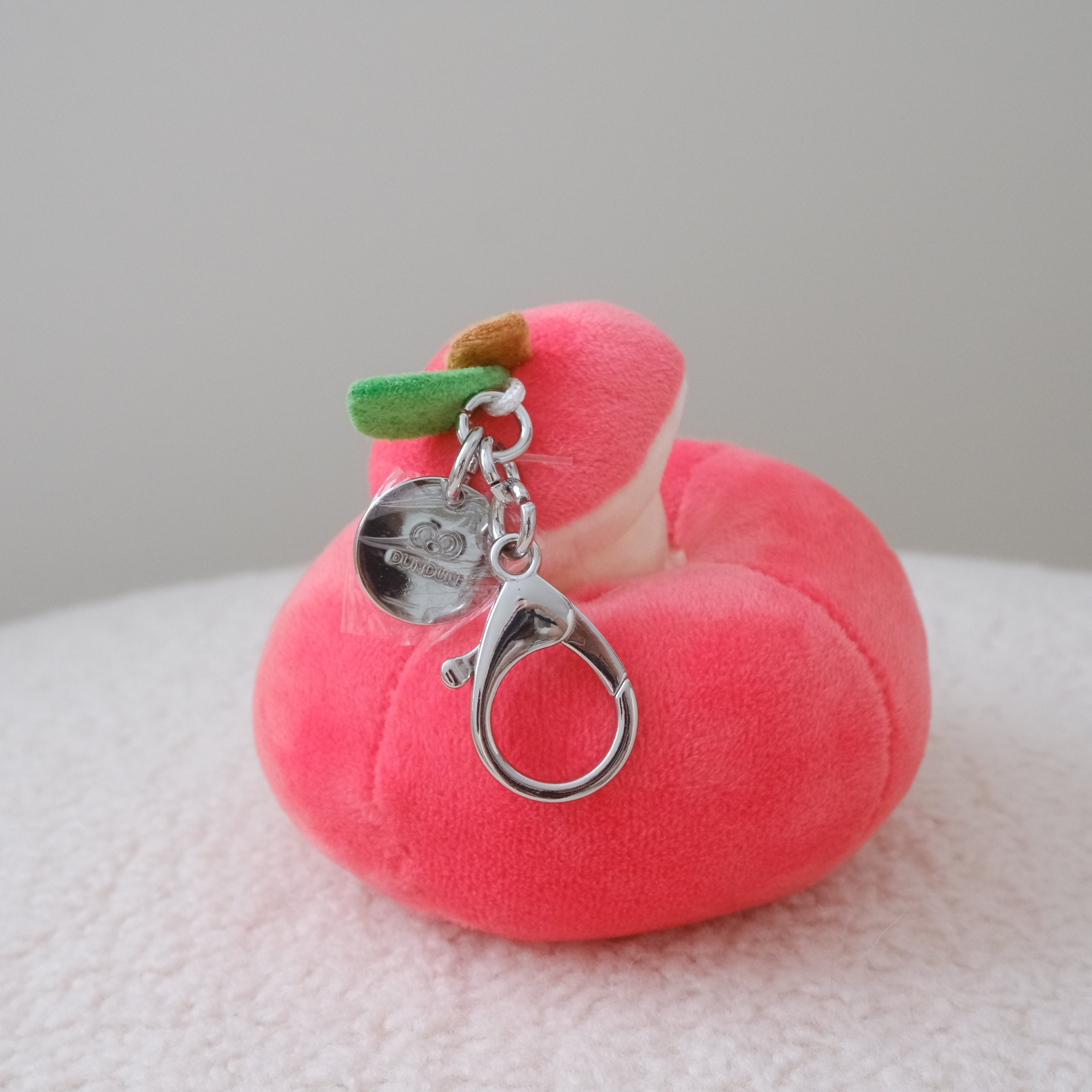 A small plush apple-shaped keychain in bright red with a silver clasp and metal charm, resting on a white textured surface against a light gray background.