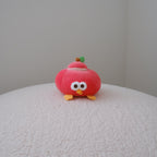 A small red plush toy shaped like an apply with large white eyes, yellow feet, and a tiny green leaf on top, sitting on a textured white surface against a plain light gray wall.