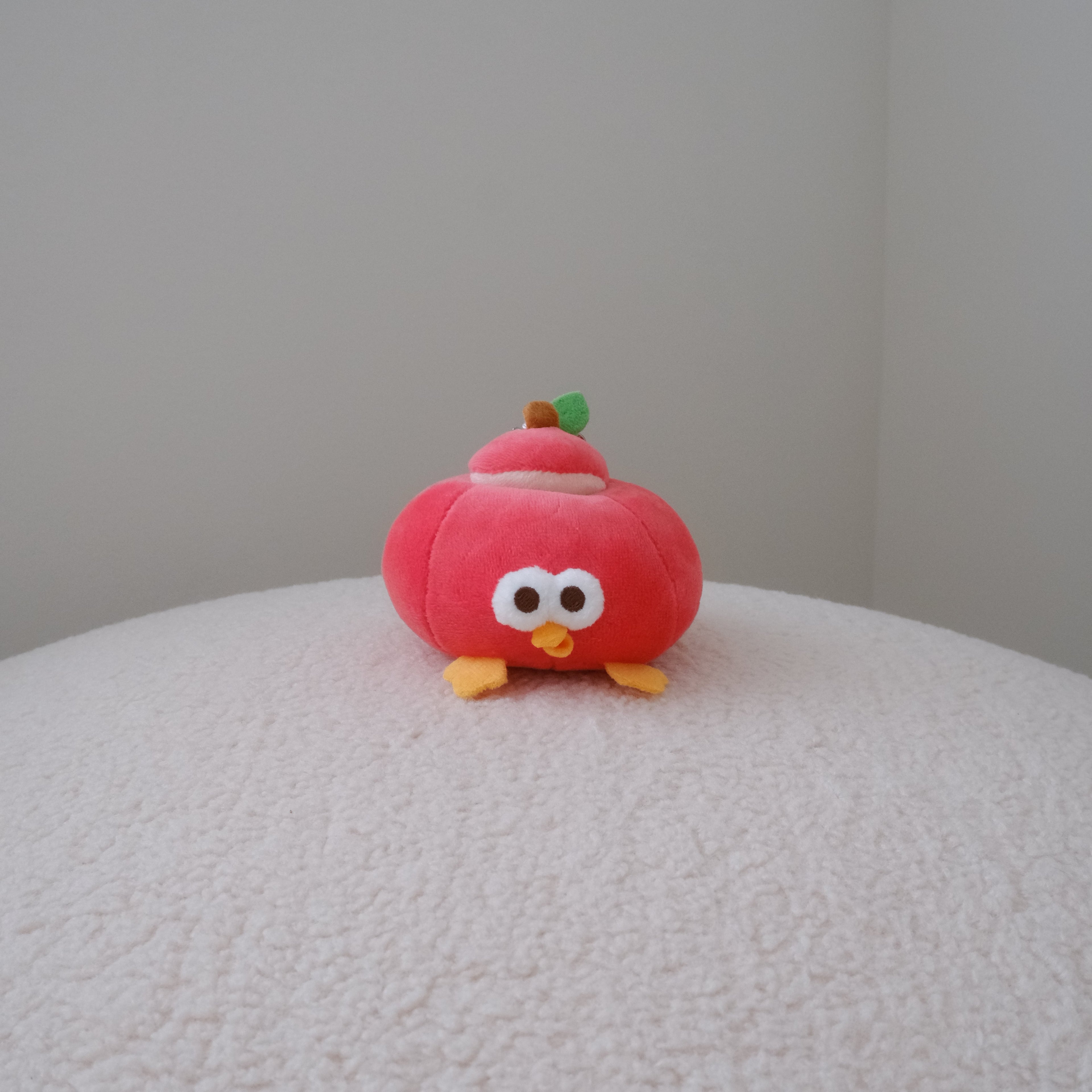 A small red plush toy shaped like an apply with large white eyes, yellow feet, and a tiny green leaf on top, sitting on a textured white surface against a plain light gray wall.