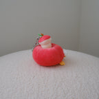 A small plush keychain shaped like a red apple with a white inner core and tiny orange feet, resting on a textured white surface against a plain light gray wall.
