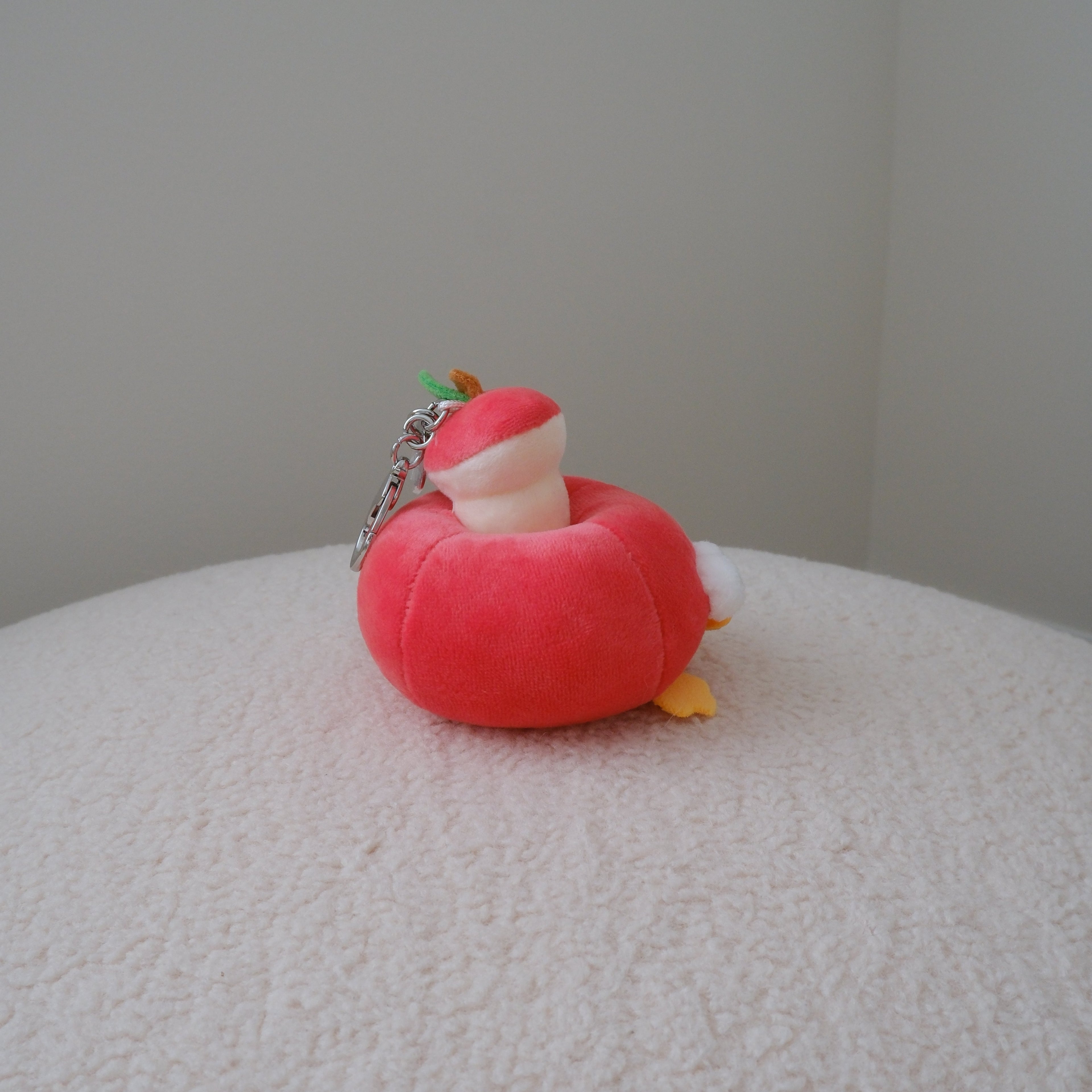 A small plush keychain shaped like a red apple with a white inner core and tiny orange feet, resting on a textured white surface against a plain light gray wall.
