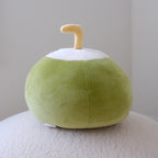 A plush coconut-shaped plush in light green and white with a small yellow stem, resting on a textured white surface against a plain light gray wall.