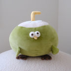 A cute green plush toy shaped like a coconut bird with a yellow straw, white top, and brown feet, sitting on a textured white surface against a plain light gray background.