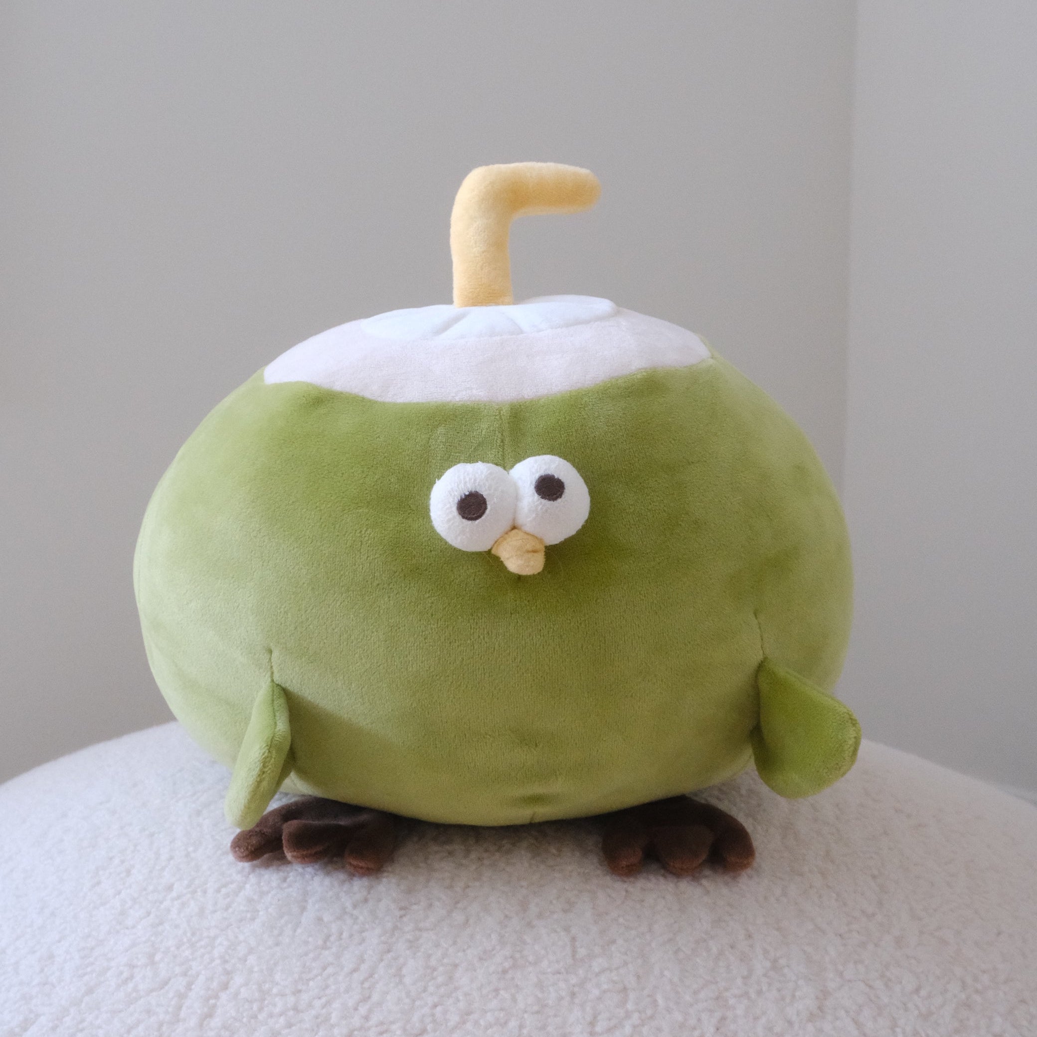 A cute green plush toy shaped like a coconut bird with a yellow straw, white top, and brown feet, sitting on a textured white surface against a plain light gray background.