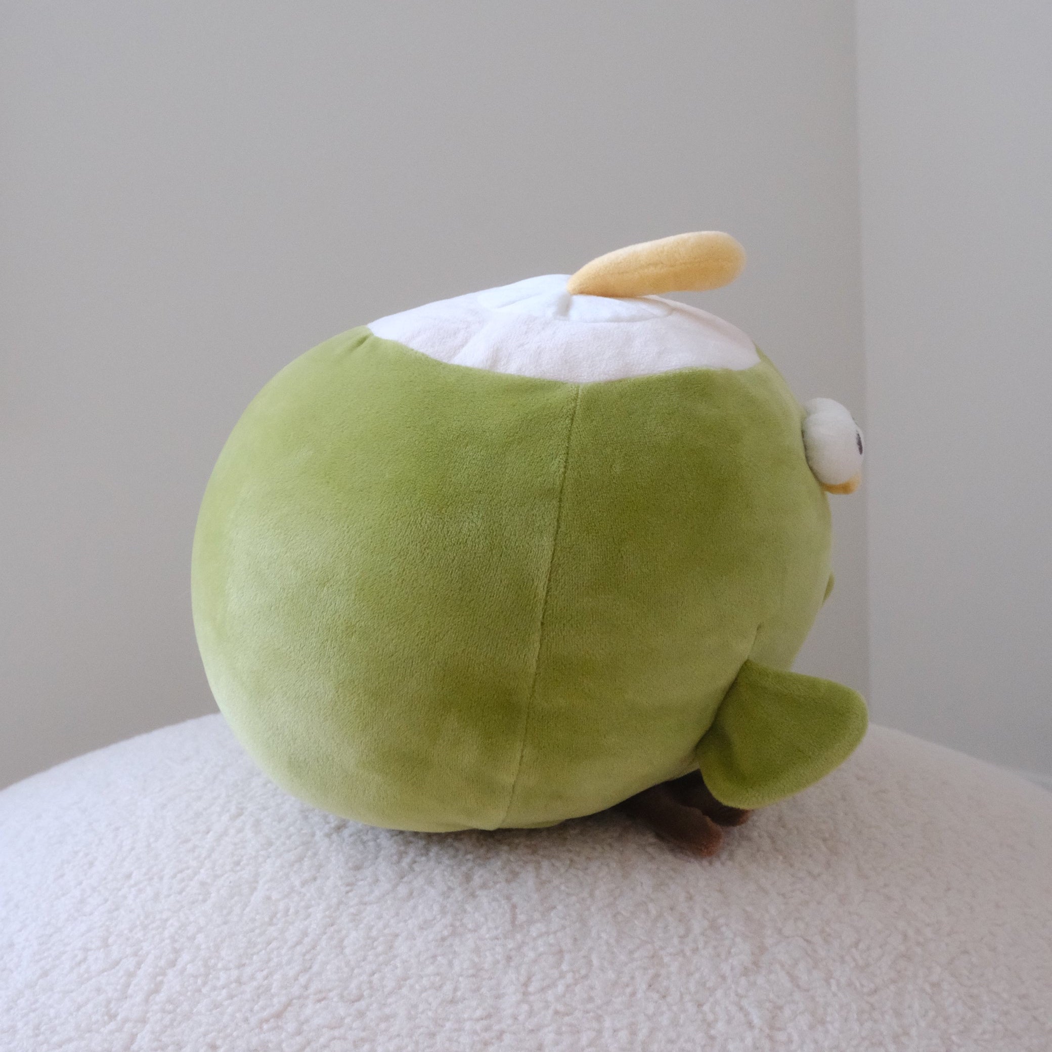 A green plush toy with a white top section and a small yellow accent, photographed sitting on a textured white surface against a plain light gray wall.
