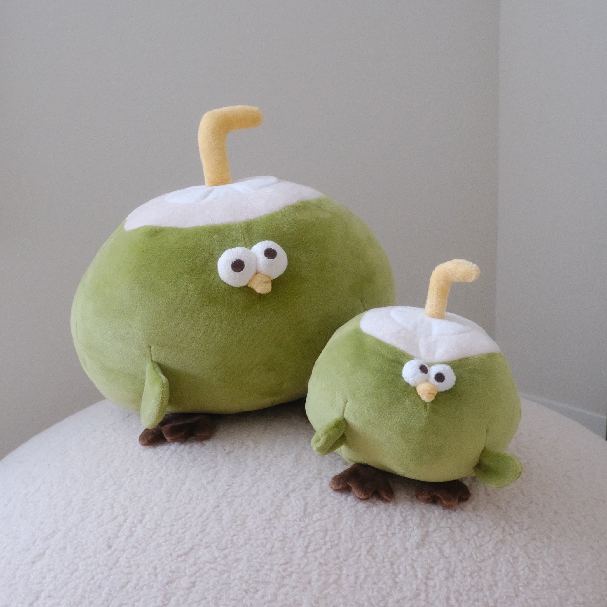 Two cute green coconut-shaped plush toys with white tops and yellow straw-like handles, sitting on a white textured surface against a plain light gray wall.
