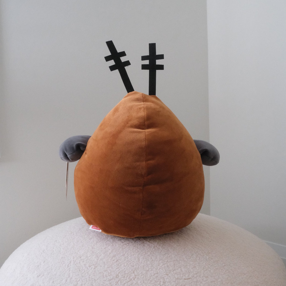 A brown plush toy with black antler-like details and small gray ears, sitting on a round white textured cushion against a plain light gray wall.