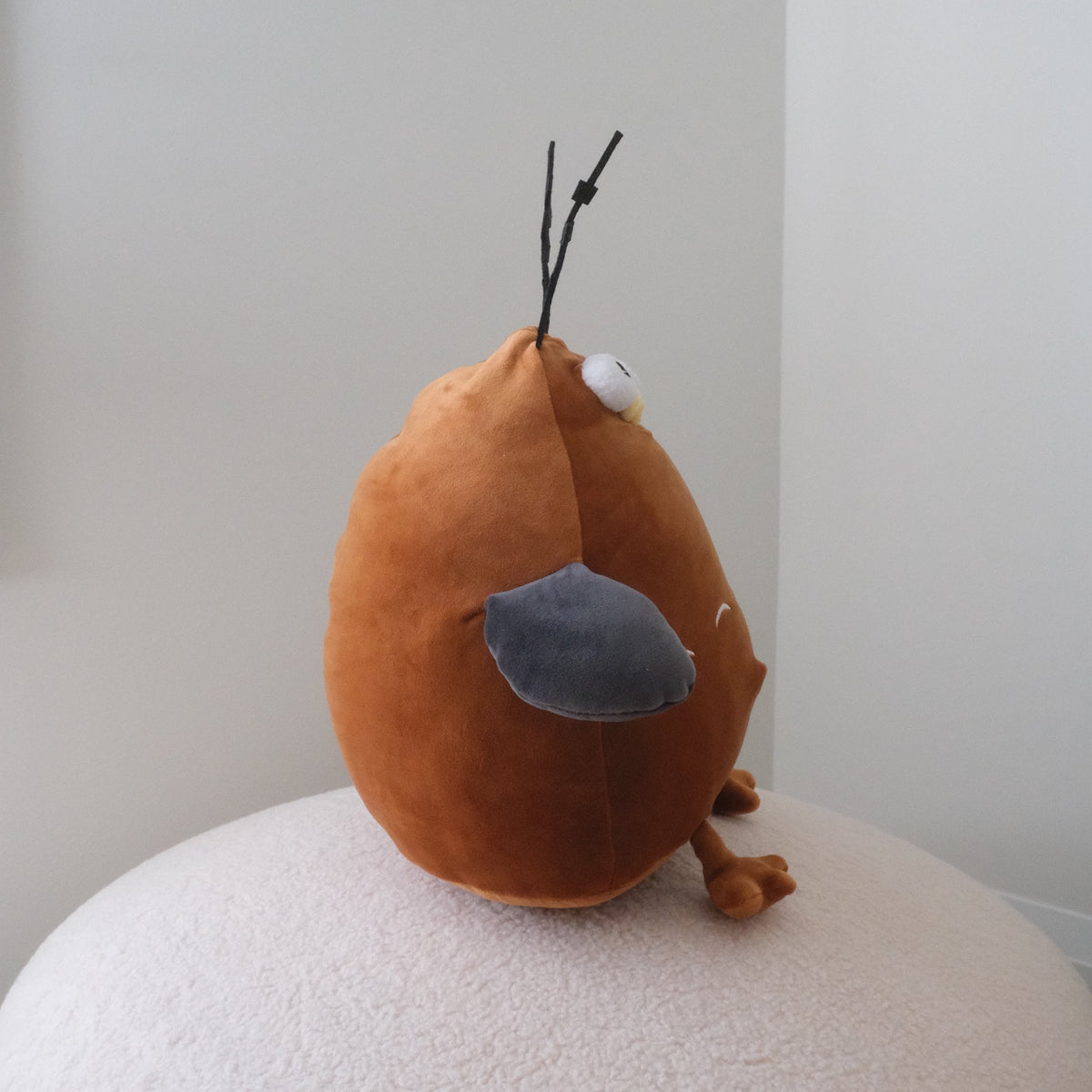 A brown plush toy shaped like a bird with a gray beak and black antennae, sitting on a round white textured cushion against a plain light gray wall.
