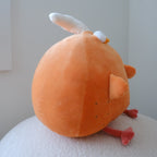 An orange plush toy with a white bone on its head and small red feet, photographed from the back while sitting on a textured white surface against a plain light gray wall.