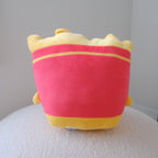 A plush toy shaped like a French fries bag, featuring a bright red body with yellow trim and a yellow top, sitting on a textured white surface against a plain light gray background.
