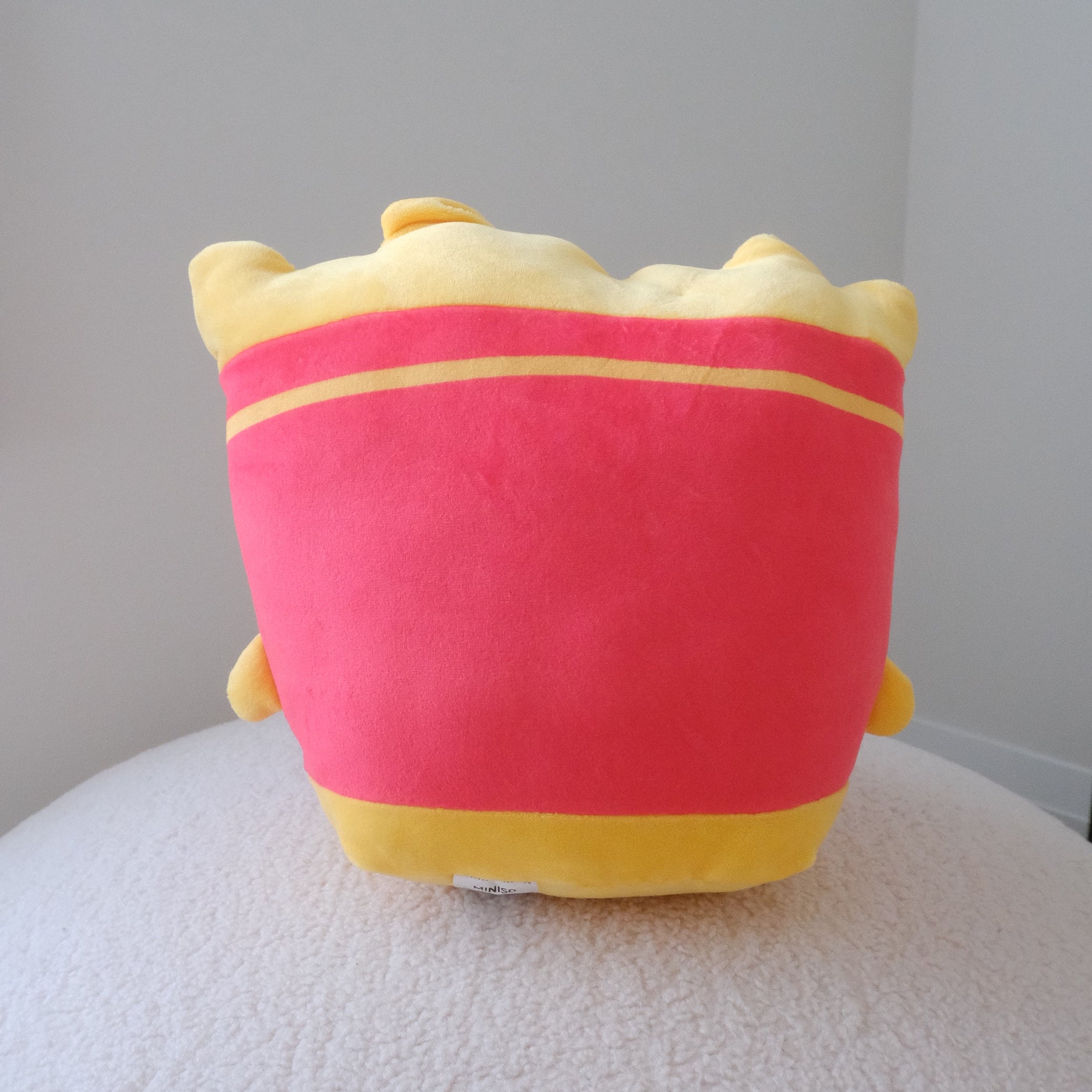 A plush toy shaped like a French fries bag, featuring a bright red body with yellow trim and a yellow top, sitting on a textured white surface against a plain light gray background.