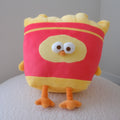 A plush toy shaped like a yellow and red french fries bag with cartoon eyes and orange feet, sitting on a white textured surface against a plain light gray wall.

