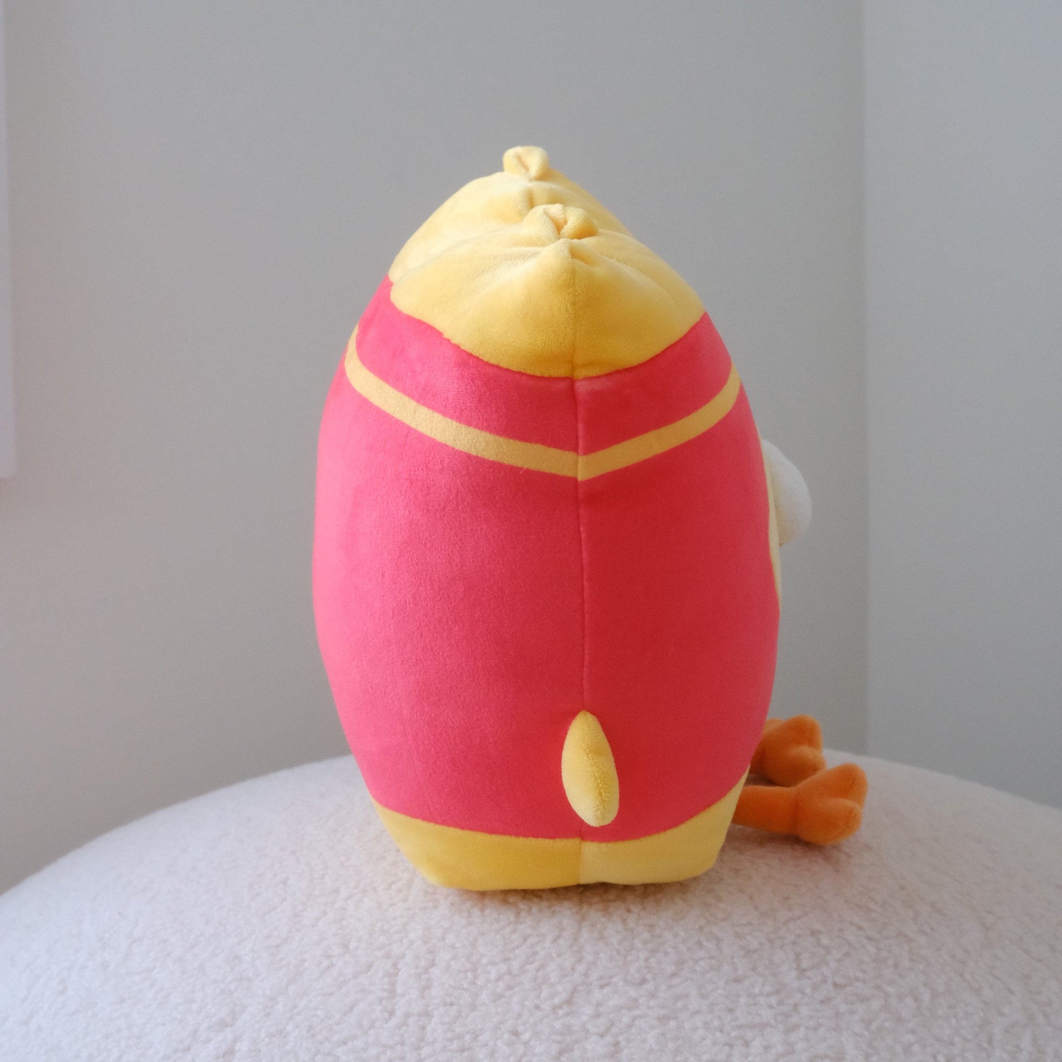 A plush toy shaped like french fries, shown from the side while sitting on a textured white surface against a plain light gray wall.