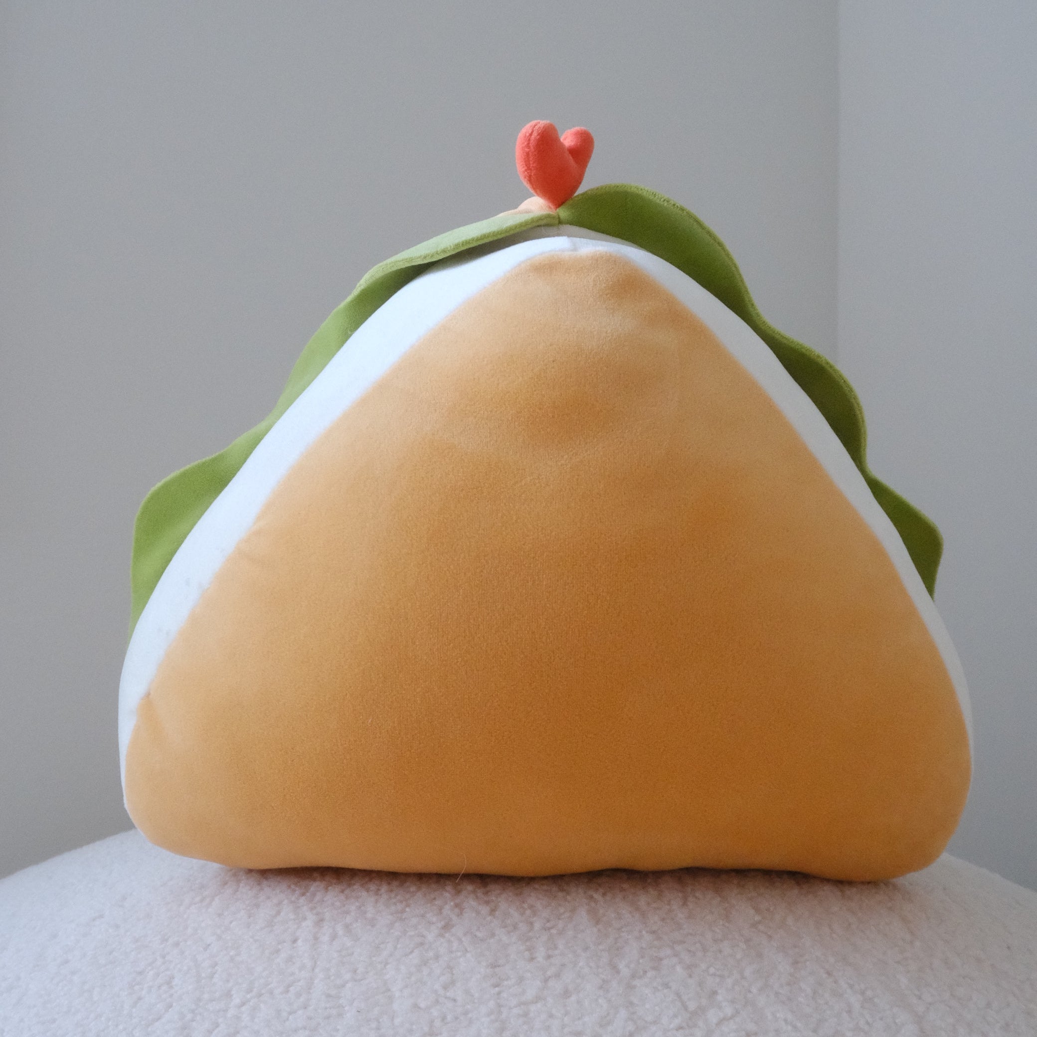 A soft plush toy shaped like a triangular sandwich with a soft yellow-orange bun, green lettuce, and a small red heart on top, resting on a textured white surface against a neutral gray background.