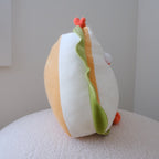 A plush toy shaped like a sandwich, featuring white and yellow buns, green lettuce, and orange tomato pieces, sitting on a textured white surface against a neutral light gray background.