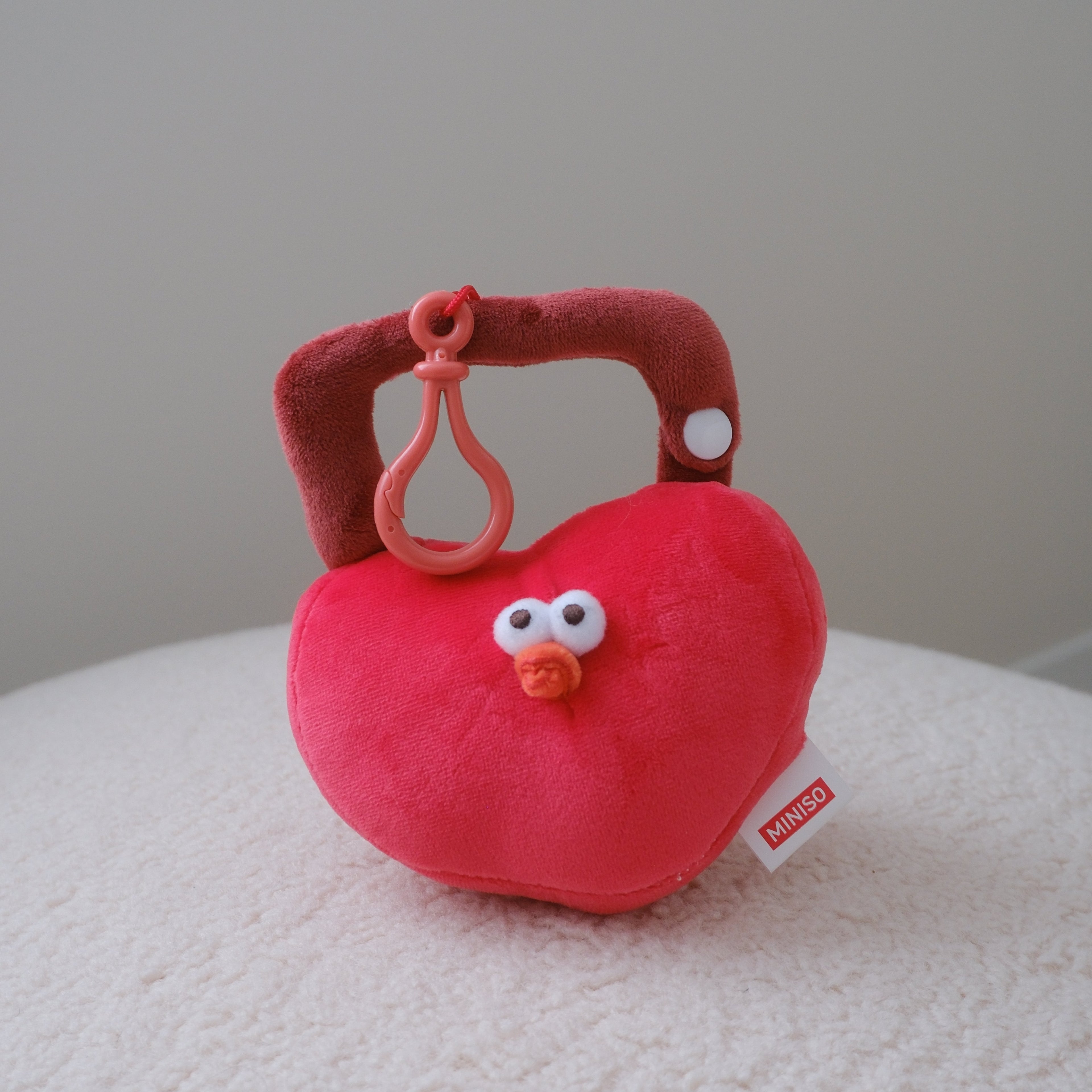 A red plush heart-shaped lock resting on a white textured surface against a neutral gray background. A 'MINISO' brand tag is visible on the side.