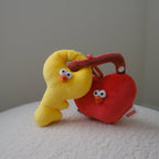 A yellow plush key and a red plush heart-shaped lock, both featuring cartoon eyes and orange beaks, posed together on a white textured surface against a neutral gray background.